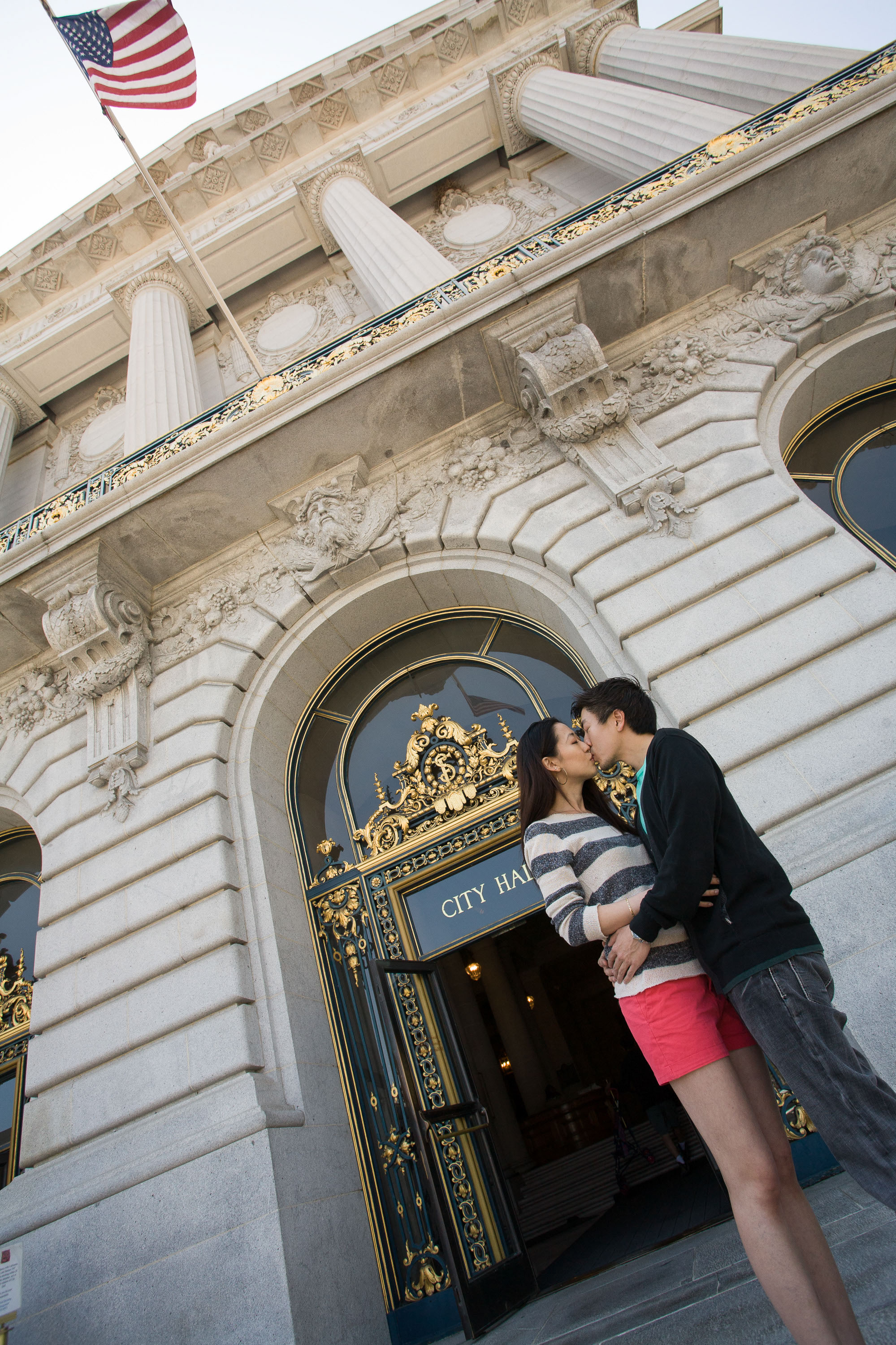 A kiss framed by a dramatic view of the columns and filigree, and the American Flag, at a San Francisco City Hall wedding
